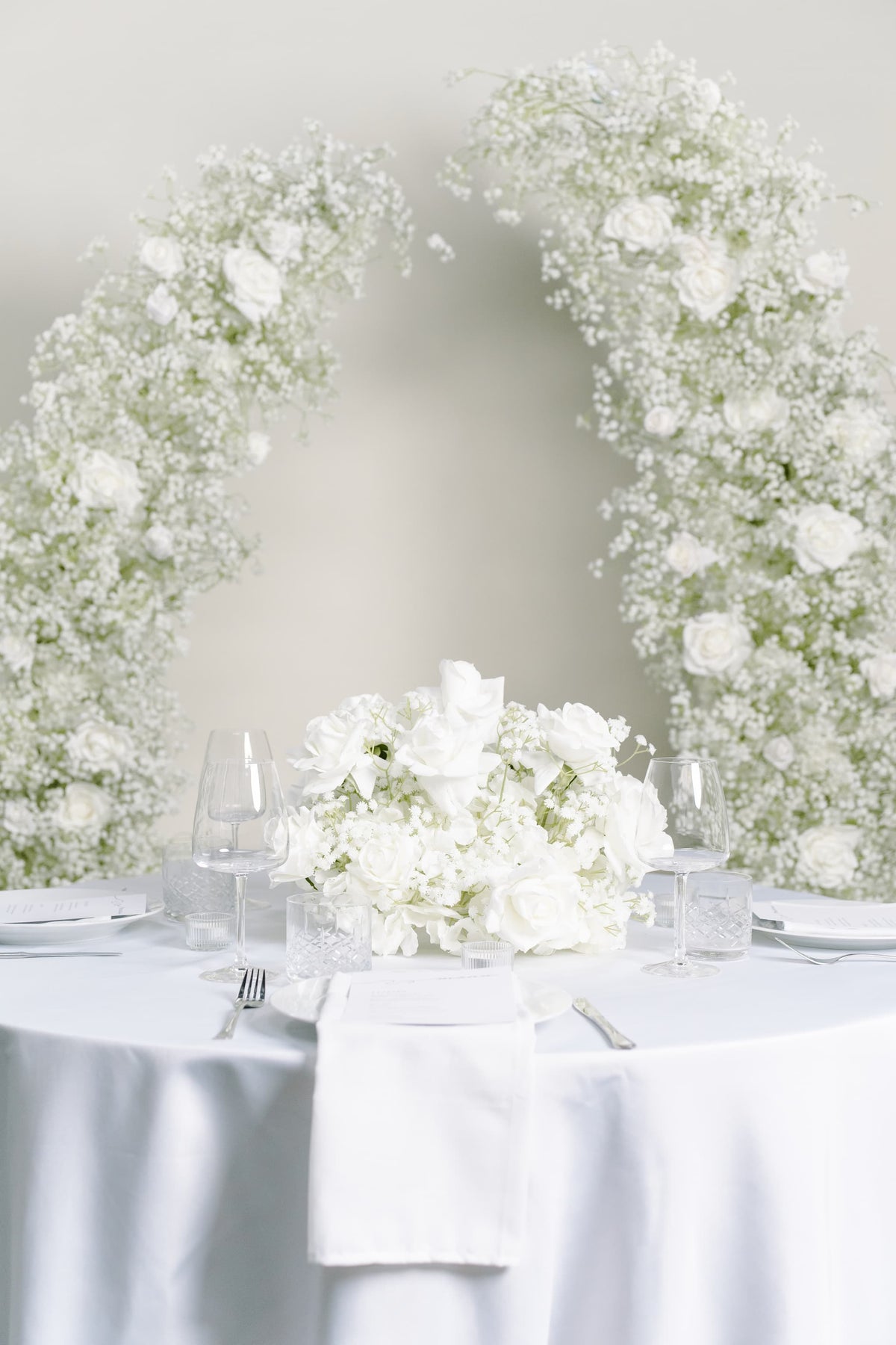 A round white table set with a central bouquet of white roses, clear wine glasses, folded white napkins, and silver cutlery, with a large floral arch made of white roses and baby's breath behind it.