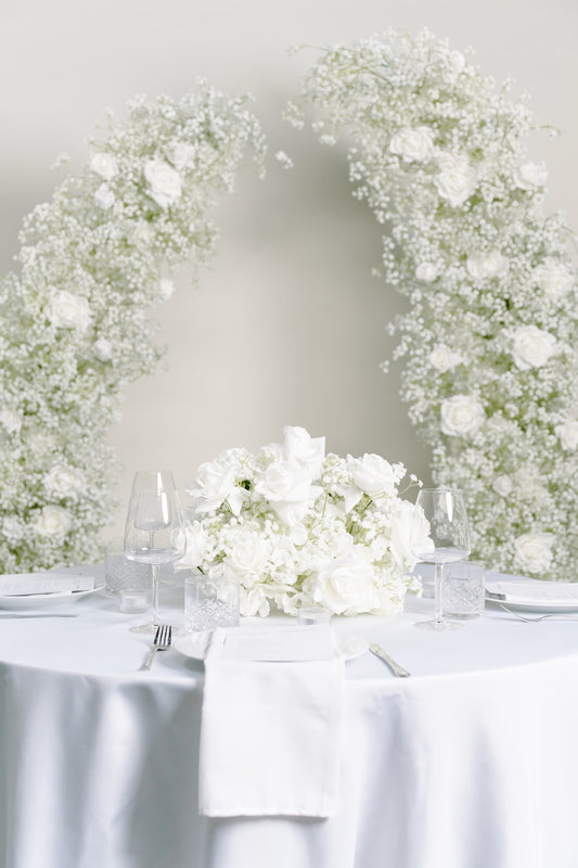 A round white table set with a central bouquet of white roses, clear wine glasses, folded white napkins, and silver cutlery, with a large floral arch made of white roses and baby's breath behind it.